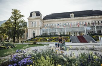 Eine Frau und ein Mann sitzen am Rand eines Brunnens im Park des Theater u. Kongresshauses Bad Ischl im Salzkammergut.