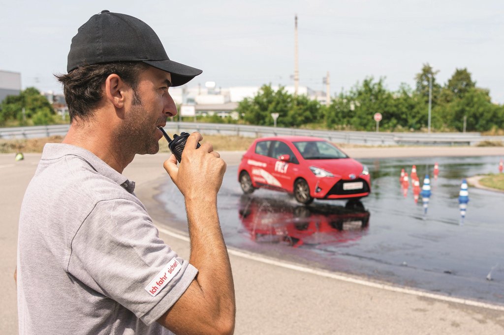 Auf dem Foto sieht man den Trainer im Vordergrund während hinter ihm ein Fahrzeug auf nasser Fahrbahn fährt.